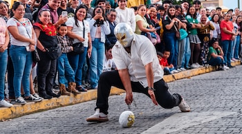 Hombre con máscara plateada inclinado sobre una calle de piedra rodeado por personas que observan en la acera.