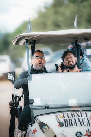 Carrito de golf blanco, dos hombres, gafas de sol, cámara fotográfica, calcomanías de Fernet Branca, bandera con texto