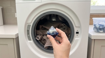 Mano que sostiene una cápsula de detergente azul y blanco frente a una lavadora blanca abierta con ropa en su interior.