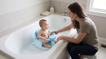Una mujer sentada junto a una bañera observa a un bebé ubicado dentro de un asiento de plástico azul con agua.
