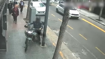 Un motociclista con el rostro difuminado circula por la acera de una calle donde transitan peatones y automóviles