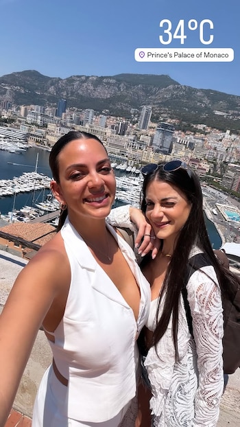 Dos mujeres sonriendo en primer plano, con vista panorámica de Mónaco, sus edificios, puerto con yates y montañas. Se muestra 34 grados Celsius y Palacio del Príncipe