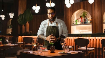 Un chef barbudo con gafas y delantal verde se alza en un restaurante con luz tenue, sosteniendo una planta pequeña sobre una mesa con copas y platos