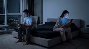 Hombre y mujer sentados en lados opuestos de una cama en la oscuridad, ambos mirando sus teléfonos móviles cuyas pantallas iluminan sus rostros.