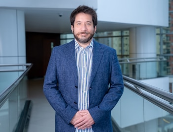 Un hombre con barba y saco azul posa con las manos entrelazadas en un pasillo moderno con barandilla de vidrio
