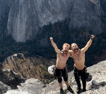 Dos hombres con torso desnudo, arneses y cascos, celebran sobre una roca con los brazos levantados frente a una gran montaña y un valle arbolado