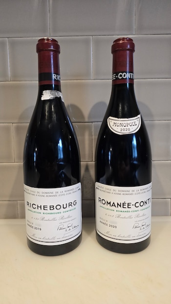 Dos botellas de vino de vidrio oscuro con etiquetas blancas detalladas y tapones rojos se posan sobre una superficie clara frente a una pared de azulejos grises