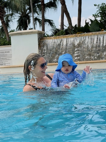 Una mujer con gafas de sol sostiene a un bebé con gorro azul dentro de una piscina, con una cascada y palmeras detrás