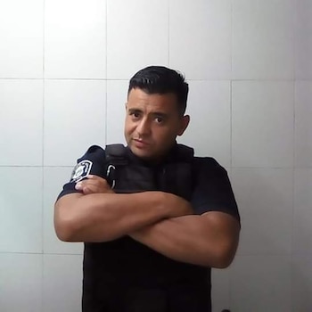 Hombre de cabello oscuro, con camisa negra y chaleco antibalas, mira a la cámara con los brazos cruzados. Fondo de pared de azulejos blancos