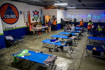 Interior de un albergue de Conred con catres azules alineados, personas sentadas y de pie, y el logotipo de Conred en la pared