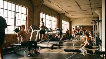 Múltiples personas se ejercitan en un gimnasio con grandes ventanales y paredes de ladrillo; se ven cintas de correr, bicicletas estáticas y pesas en uso.