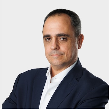Un hombre con cabello corto y oscuro viste un saco azul marino y una camisa blanca sobre un fondo gris claro