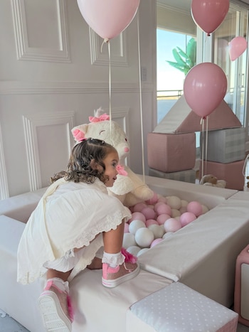Una niña con vestido blanco y zapatillas rosadas se apoya sobre un pelotero con pelotas rosadas y blancas junto a varios globos