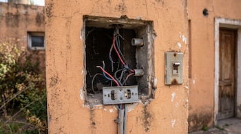 Un hueco en una pared de color beige contiene cables eléctricos y tubos de protección sin un medidor, junto a un interruptor de luz.