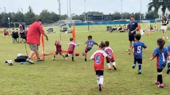 Varios niños con uniformes deportivos corren hacia una portería pequeña sobre un campo de césped mientras un adulto observa la jugada