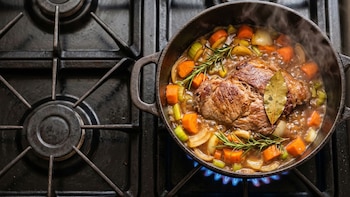 Cacerola de hierro fundido sobre una hornalla de gas azul encendida. Contiene bondiola de cerdo, zanahorias, cebollas, apio, romero y laurel burbujeando