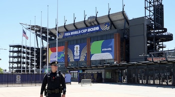 Oficial de policía con uniforme, gorra y cámara corporal frente a estadio. Banderas de la Copa Mundial FIFA 2026. Bandera de Estados Unidos.
