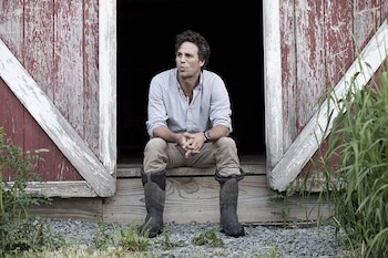 Mark Ruffalo sentado en el umbral de un granero de madera con camisa clara, pantalón caqui y botas altas