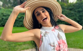 Una mujer de cabello oscuro y sonrisa amplia, con un sombrero de paja y un vestido blanco floral, posa feliz al aire libre con fondo verde