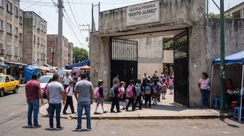Vista exterior de la Escuela Primaria "Benito Juárez" en la Ciudad de México, con su portón abierto por donde entran niños con mochilas, y adultos observando.