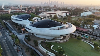 Edificio blanco y curvo del Lucas Museum of Narrative Art con techos verdes y paneles solares, rodeado de césped, una carretera y el horizonte de Los Ángeles