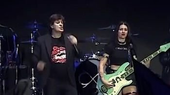 Dos músicos en un escenario oscuro. A la izquierda, una persona canta con micrófono. A la derecha, una mujer toca una guitarra eléctrica verde claro