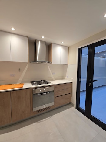 Cocina moderna con gabinetes blancos y de madera clara, encimera blanca, campana extractora y horno de acero inoxidable. Puerta de vidrio a la derecha