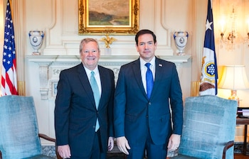 Marco Rubio y el jefe de la misión estadounidense posan de traje frente a una chimenea flanqueados por banderas de Estados Unidos
