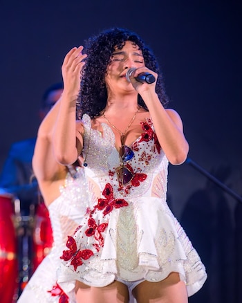 Mujer con cabello rizado, micrófono, ojos cerrados, mano alzada. Viste vestido blanco brillante con diseño de mariposas rojas. Músico al fondo