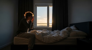 Persona sentada en una cama con sábanas blancas se frota los ojos al amanecer, con luz solar tenue entrando por una ventana en un dormitorio oscuro.