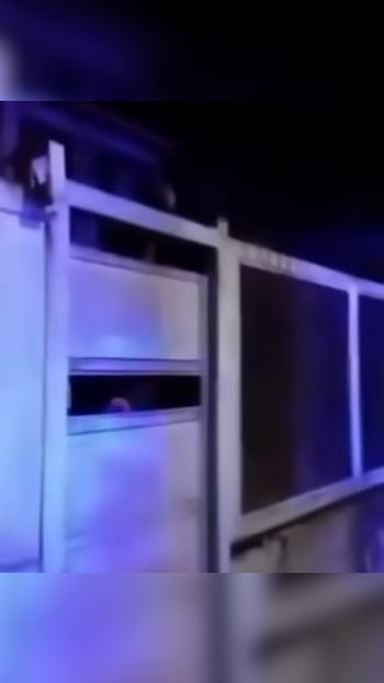 Una reja metálica de color claro con una persona visible a través de una abertura horizontal en la oscuridad bajo una luz azul
