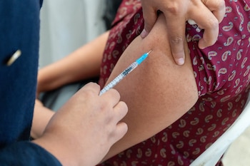 Una jeringa se introduce en el brazo de una persona. Dos manos sujetan la jeringa y el brazo. Se observa una manga de tela con estampado