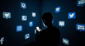 Persona de espaldas en habitación oscura usa celular. Íconos de redes sociales como Facebook, Instagram, Twitter, chat y corazón se proyectan en las paredes.