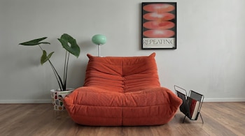 Sillón Toga naranja de pana en primer plano, con una planta en maceta decorada, una lámpara verde claro y un póster enmarcado sobre una pared blanca.