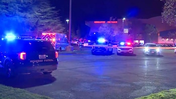 Patrullas policiales, una ambulancia y cinta perimetral en un estacionamiento nocturno frente a un edificio con letrero iluminado