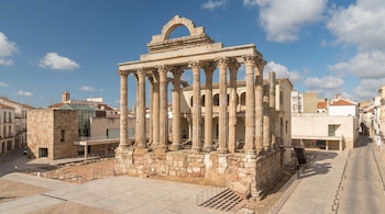 Templo romano de piedra con columnas corintias y un arco superior, situado en una plaza peatonal rodeada de edificios modernos y calles.