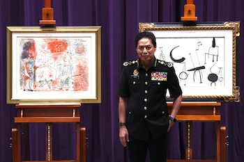 Un oficial malasio con uniforme oscuro y medallas posa entre dos obras de arte: un Picasso a la izquierda y un Miró a la derecha, sobre fondo morado