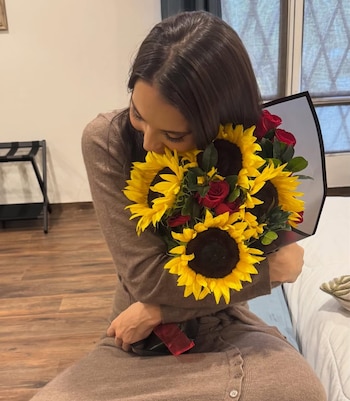 Majo Aguilar, de cabello castaño, abraza un ramo grande de girasoles amarillos y rosas rojas. Viste un suéter y falda marrones