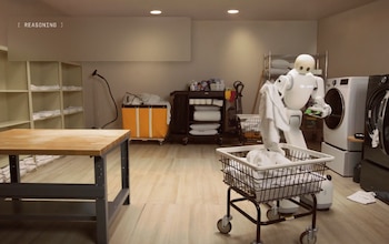 Un robot humanoide blanco toma una toalla junto a un carro con cesto, lavadoras, estantes y una mesa de madera