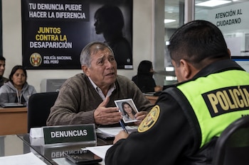 Un hombre sentado frente a un policía sostiene la fotografía de una mujer frente a un letrero con la palabra Denuncias