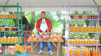Un hombre sonríe detrás de un puesto de madera que exhibe duraznos frescos, botellas de jugo y estantes con frascos de conservas. Hay flores en un balde azul.