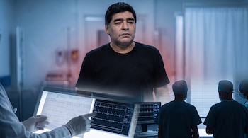 Primer plano de Diego Maradona con camiseta negra. Manos con guantes médicos señalan un estudio de electrocardiograma en un monitor. Tres médicos observan una pantalla clínica.