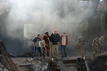 Cuatro civiles y dos soldados con fusiles sobre escombros. Hay humo y restos de edificios. Un hombre sostiene una consola con componentes electrónicos