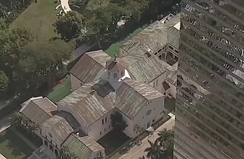 Vista aérea de una iglesia con tejados de tejas y cúpula central junto a un rascacielos de cristal en un entorno urbano con árboles y una calle