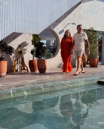 Hombre y mujer adultos caminan junto a una piscina. Ella con vestido naranja y él con camiseta clara y bermudas. Fondo de edificio de piedra con plantas y ventana
