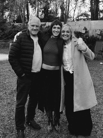 Tres personas adultas sonríen al aire libre. Un hombre y dos mujeres posan en un jardín con césped, árboles y una valla. Imagen en blanco y negro