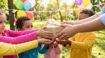 Manos de un adulto entregan una caja de regalo envuelta en papel kraft con lazo beige a cuatro pares de manos de niños. Fondo con globos y árboles.