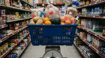 Canasta de supermercado azul con papas, naranjas, aguacate, pollo, arroz, leche, pan y pasta de dientes, sobre una balanza antigua en pasillo de supermercado con estantes y precios.