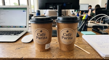 Dos vasos de café de Café Parita sobre una mesa de madera junto a una computadora portátil, un ratón inalámbrico y un cuaderno de notas.
