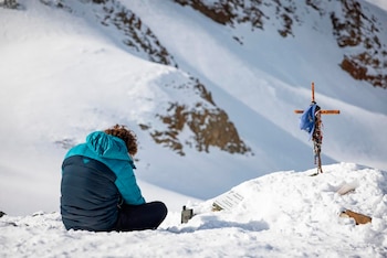 Una persona con chaqueta azul de abrigo está sentada de espaldas en la nieve junto a una placa y una cruz de madera con objetos